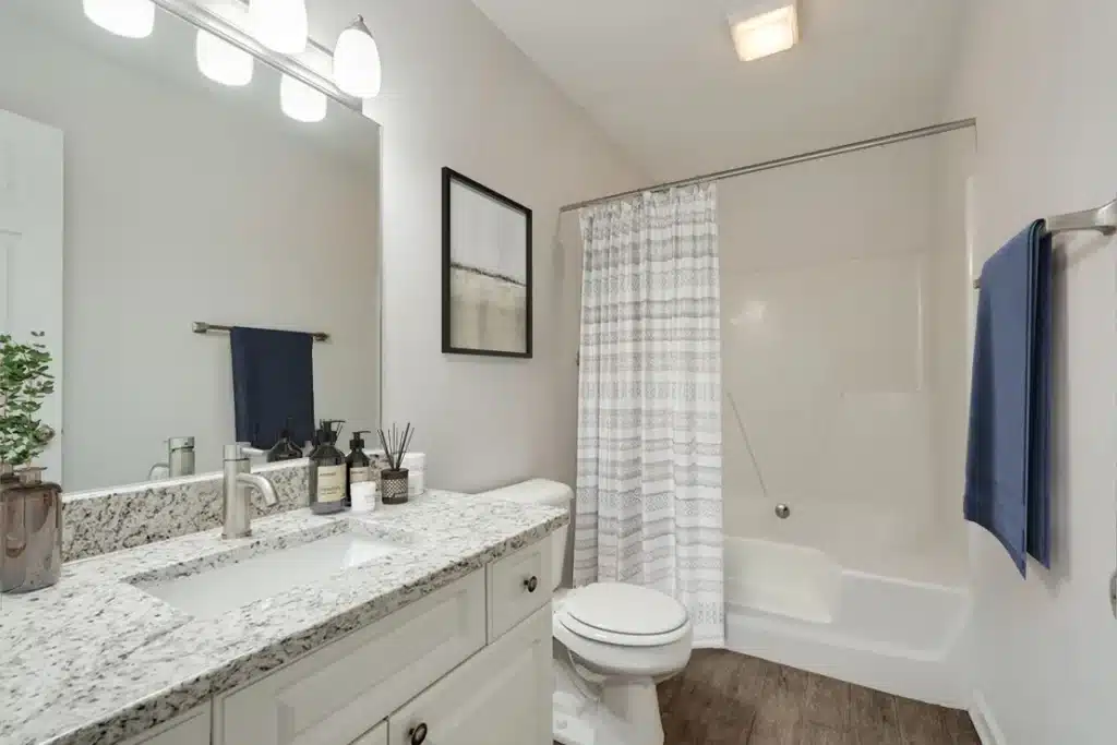 A modern bathroom with a granite countertop, white cabinets, a toilet, a bathtub with a shower curtain, a towel rack, and a wall-mounted light fixture.