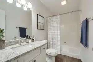 A modern bathroom with a granite countertop, white cabinets, a toilet, a bathtub with a shower curtain, a towel rack, and a wall-mounted light fixture.