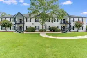 Two-story apartment building with exterior staircases on both ends, green lawn, shrubs, and a tree in front under a blue sky with scattered clouds.