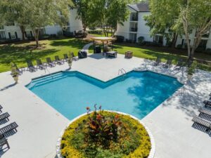 A rectangular outdoor swimming pool with poolside chairs, surrounded by trees and apartment buildings, with a circular flower bed in the foreground.