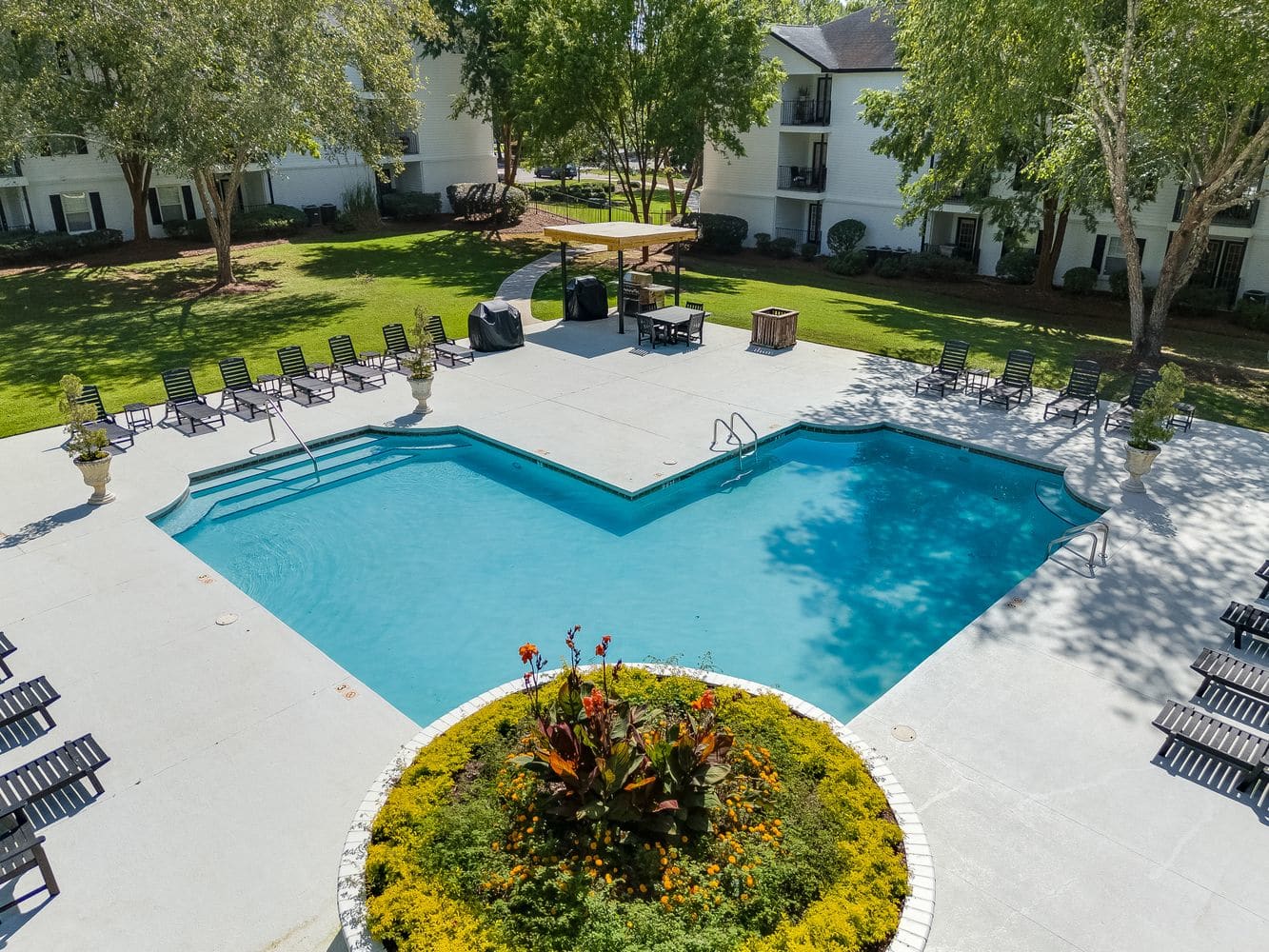 A rectangular outdoor swimming pool with poolside chairs, surrounded by trees and apartment buildings, with a circular flower bed in the foreground.