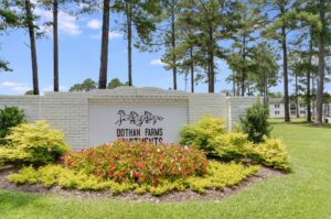 White brick sign for Dothan Farms Apartments surrounded by green shrubs and red flowers, with pine trees and apartment buildings in the background.