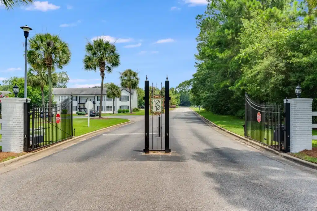 A gated driveway entrance with open black iron gates, a paved road, and residential buildings with trees and manicured lawns on both sides.