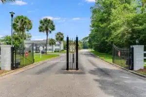A gated driveway entrance with open black iron gates, a paved road, and residential buildings with trees and manicured lawns on both sides.