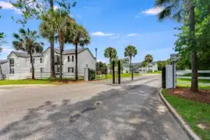 Gated entrance to a residential complex with white two-story buildings, palm trees, and a paved road under a clear sky.
