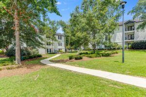 Sidewalk winding through a grassy courtyard with trees and shrubs, bordered by multi-story apartment buildings on a sunny day.
