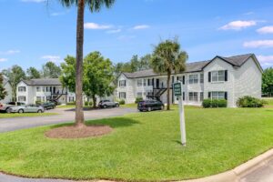 Two white, two-story apartment buildings with black roofs are seen behind a green lawn, parked cars, and a sign reading "410 Unit Drive." Trees and blue sky are in the background.