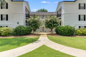 Two-story white apartment building with black shutters, surrounded by green bushes and trees, with a concrete path leading to the entrance.