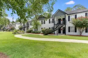 Two-story apartment building with white exterior, black shutters, and outdoor staircases, surrounded by green lawn, trees, and a curved sidewalk.