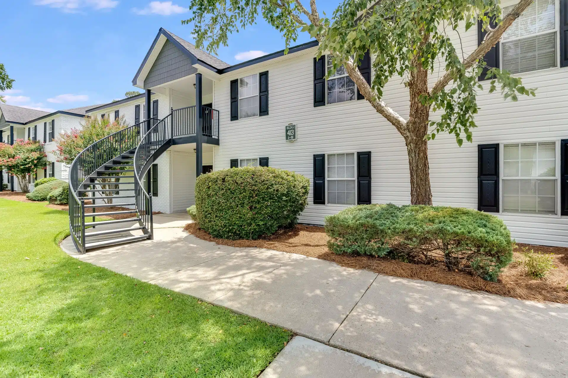 Two-story white apartment building with black shutters, an exterior staircase, and landscaped green lawn with bushes and trees.