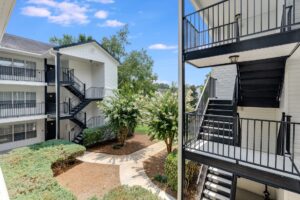View of a modern apartment complex with white buildings, black metal staircases, landscaped shrubs, trees, and a winding concrete path under a blue sky.