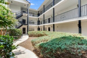 Three-story apartment building with exterior hallways, black railings, and a stairwell; landscaped bushes and a concrete walkway in the courtyard area.