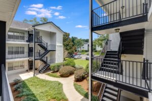 Three-story apartment buildings with exterior staircases overlook a landscaped courtyard with bushes and walkways on a sunny day.