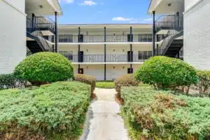 A concrete walkway bordered by trimmed bushes leads to a three-story apartment building with exterior staircases and balconies.