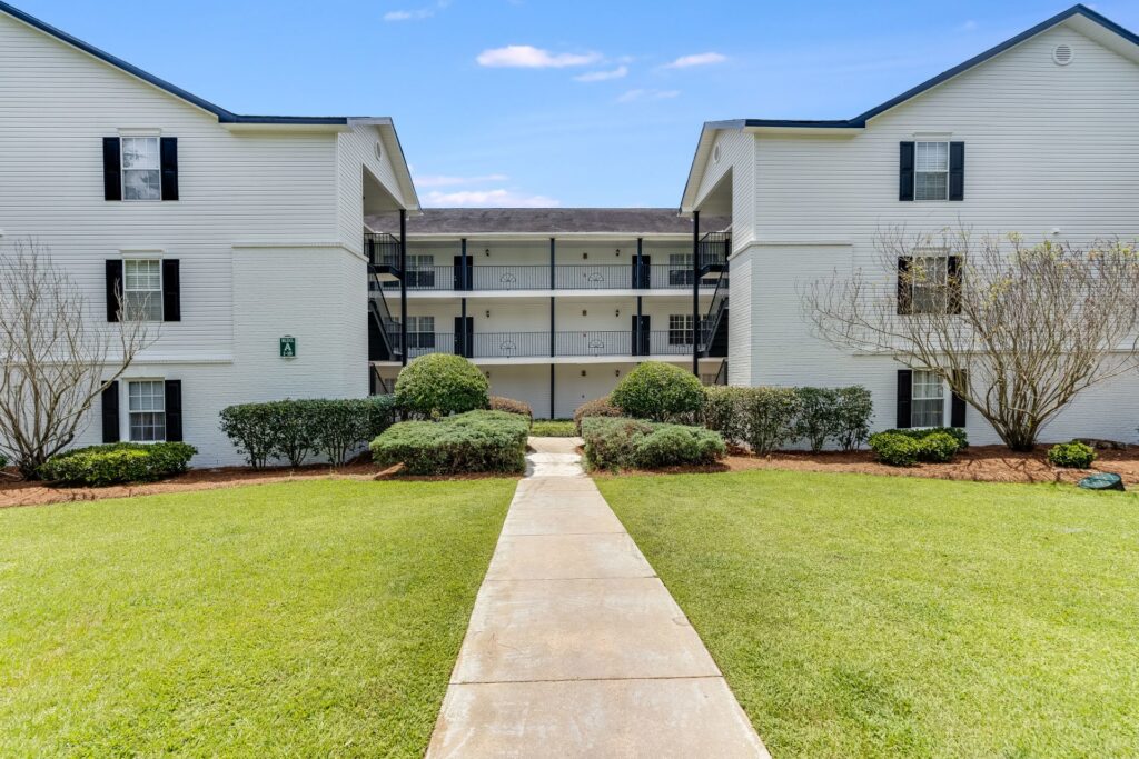 A concrete walkway leads to a three-story white apartment building with black shutters, surrounded by trimmed bushes and grass under a clear blue sky.