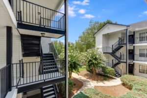 Two apartment buildings with exterior black metal staircases, light-colored siding, and landscaped paths surrounded by bushes and trees under a clear blue sky.