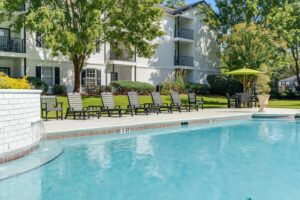 Outdoor swimming pool with lounge chairs lined up along the poolside, in front of a white apartment building with green trees and landscaping.