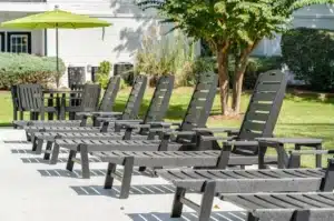 Several black lounge chairs are arranged in a row on a concrete patio outdoors, with a table and green umbrella nearby, and grass, bushes, and trees in the background.