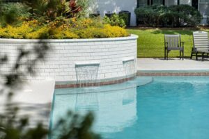 A swimming pool with clear blue water, two small waterfalls flowing from a white brick planter, and chairs on a concrete patio in the background.