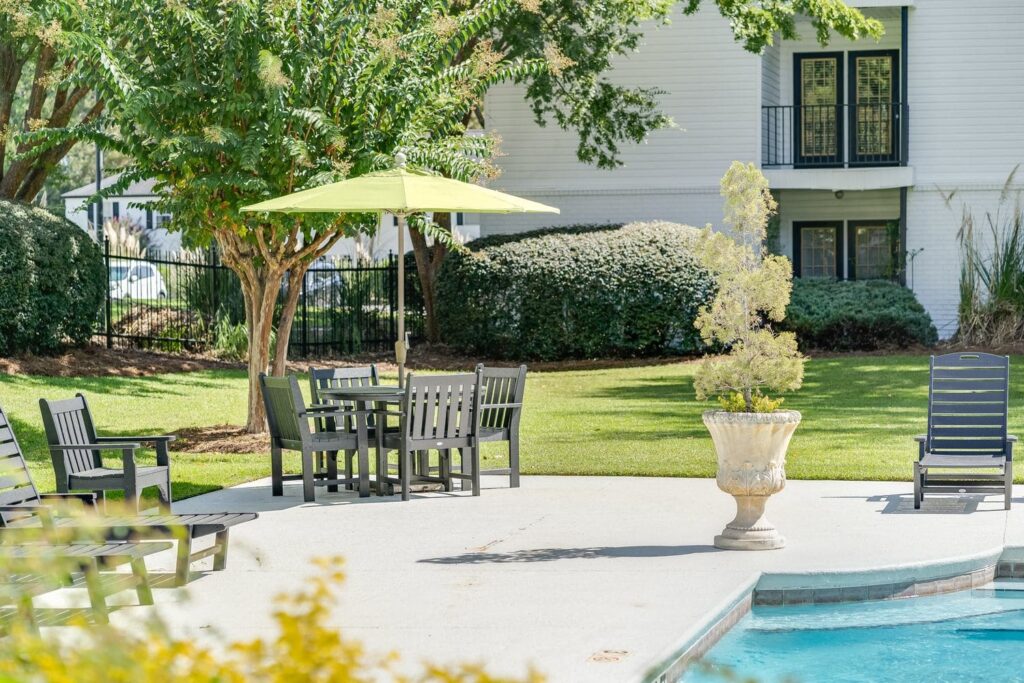 Outdoor poolside area with black lounge chairs, a table with chairs under a green umbrella, a potted plant, and landscaped greenery in the background.