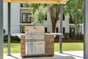 A stainless steel outdoor grill with a stone base stands under a wooden pergola in front of an apartment building and landscaped lawn.