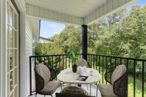 A covered balcony with a round table, four wicker chairs, coffee cups, a teapot, and a magazine overlooks a green, leafy yard.