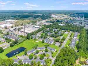 Aerial view of a suburban area with apartment buildings, roads, green lawns, a pond, and nearby commercial or industrial facilities.