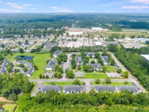 Aerial view of a residential complex surrounded by trees, roads, and parking lots, with commercial buildings and green areas in the background.