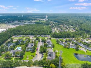 Aerial view of a suburban neighborhood with houses, parking lots, trees, a small pond, and a cell tower, surrounded by forested areas and distant roads.